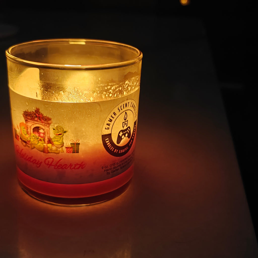 This image shows a 9 ounce Tumbler jar candle of Holiday Hearth. The candle is lit in a dark room. The candle itself is a dark magenta color. The label is a light green and features 3 cartoon orcs gathered around a holiday fireplace with gifts and holiday drinks. The side of the candle features the Gamer Scent Candles logo, which consists of a stylized candle on top of a video game controller.
