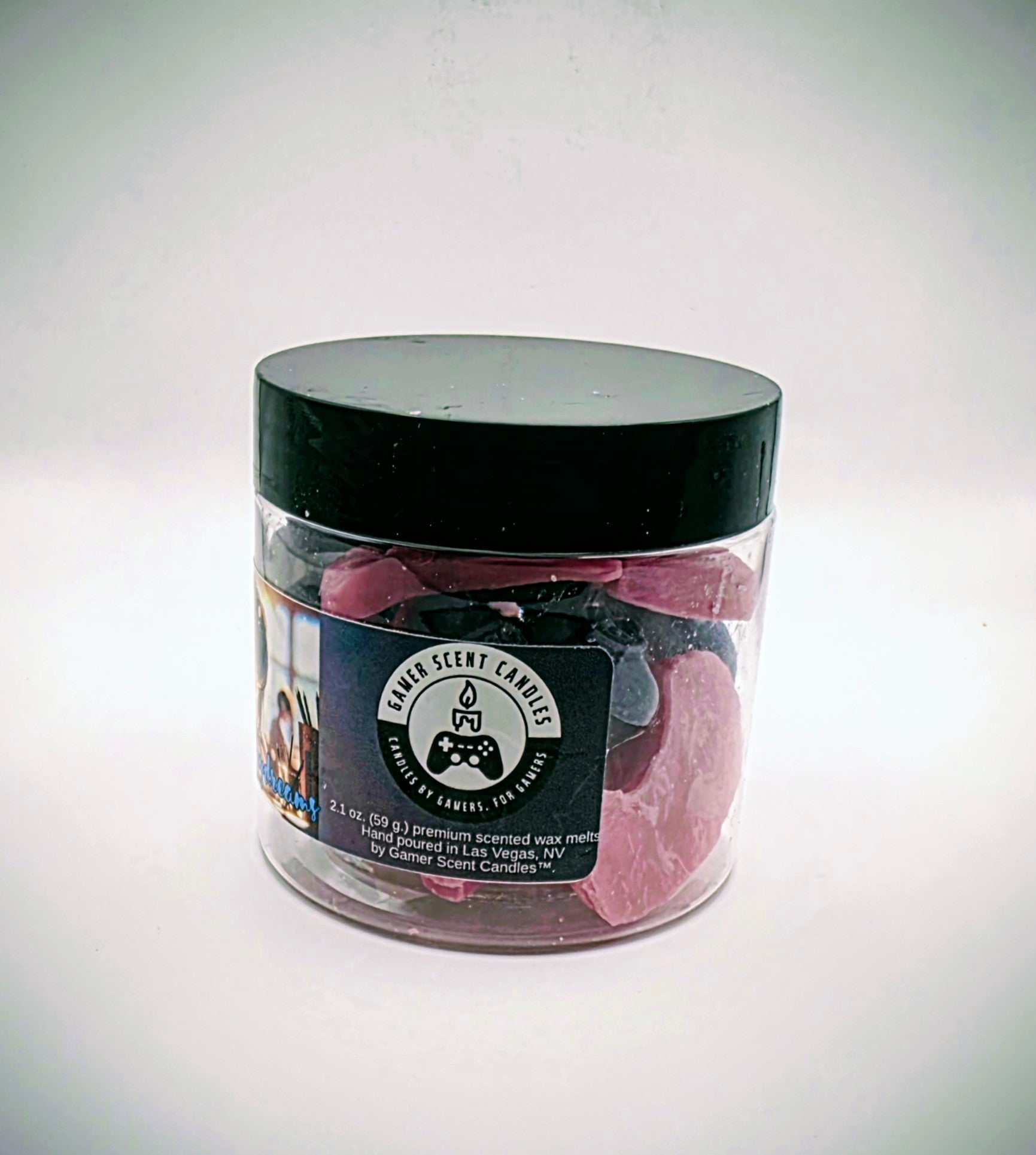 A jar of "Japanese Daydreams" premium wax melts. Each wax melt is shaped like a video game controller, and the melts come in a mixture of purple and pink. The melts come in a clear plastic jar with a black lid and feature a label of a Japanese girl at a school desk, wearing headphones, seeming to lost in thought.