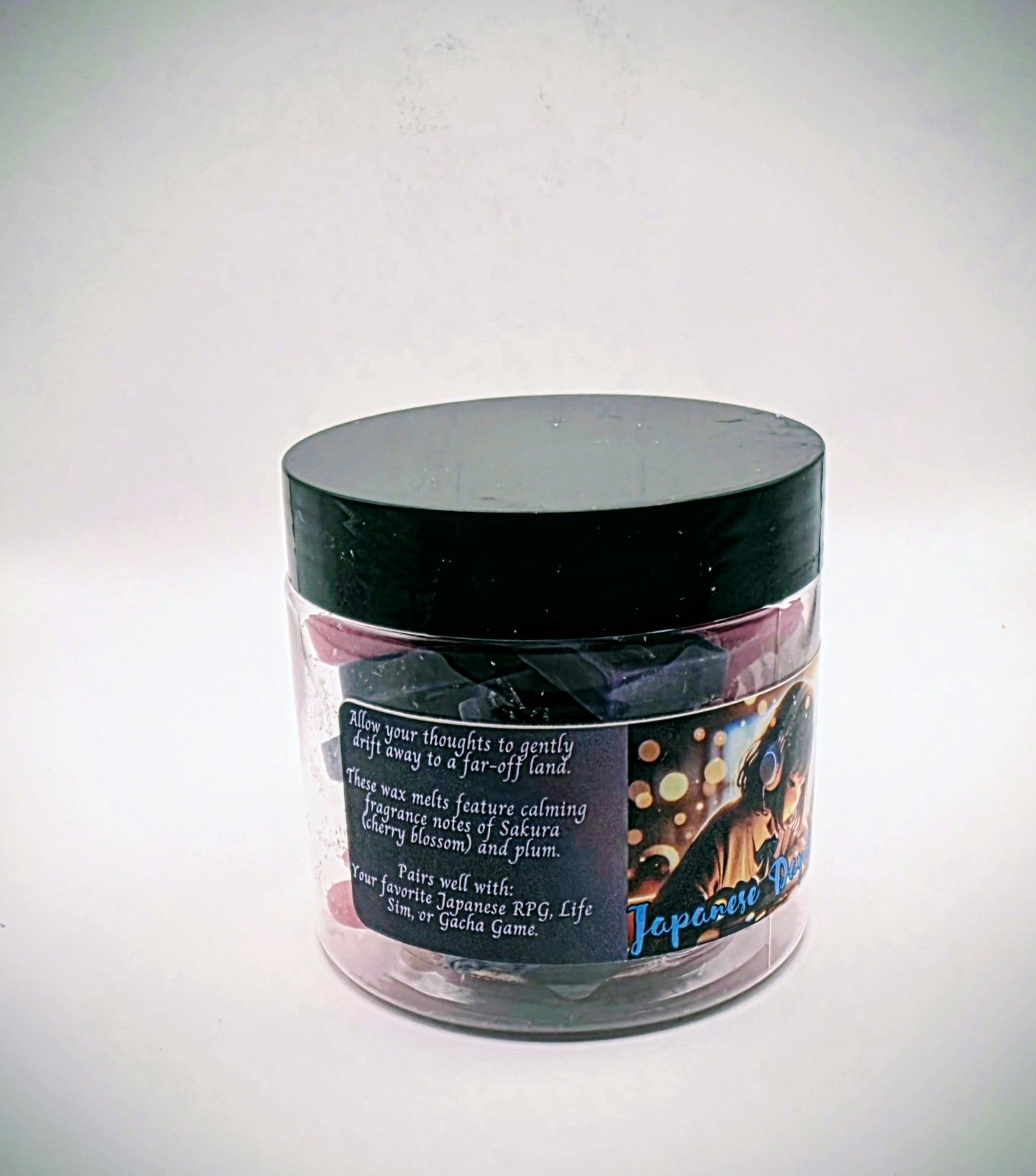 A jar of "Japanese Daydreams" premium wax melts. Each wax melt is shaped like a video game controller, and the melts come in a mixture of purple and pink. The melts come in a clear plastic jar with a black lid and feature a label of a Japanese girl at a school desk, wearing headphones, seeming to lost in thought.