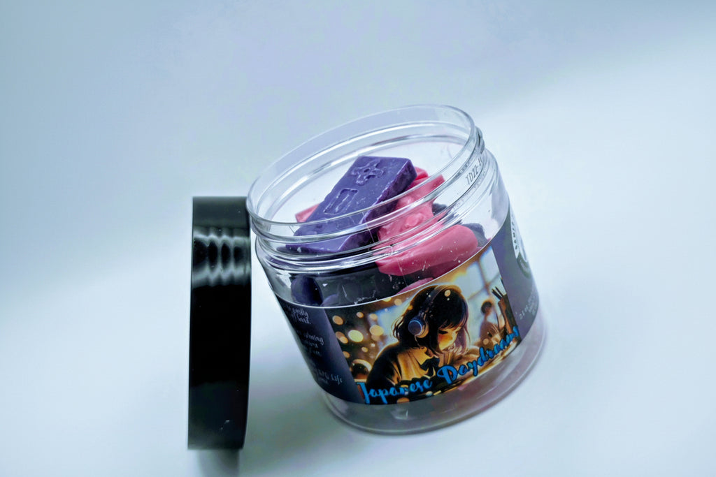 A jar of "Japanese Daydreams" premium wax melts. Each wax melt is shaped like a video game controller, and the melts come in a mixture of purple and pink. The melts come in a clear plastic jar with a black lid and feature a label of a Japanese girl at a school desk, wearing headphones, seeming to lost in thought.