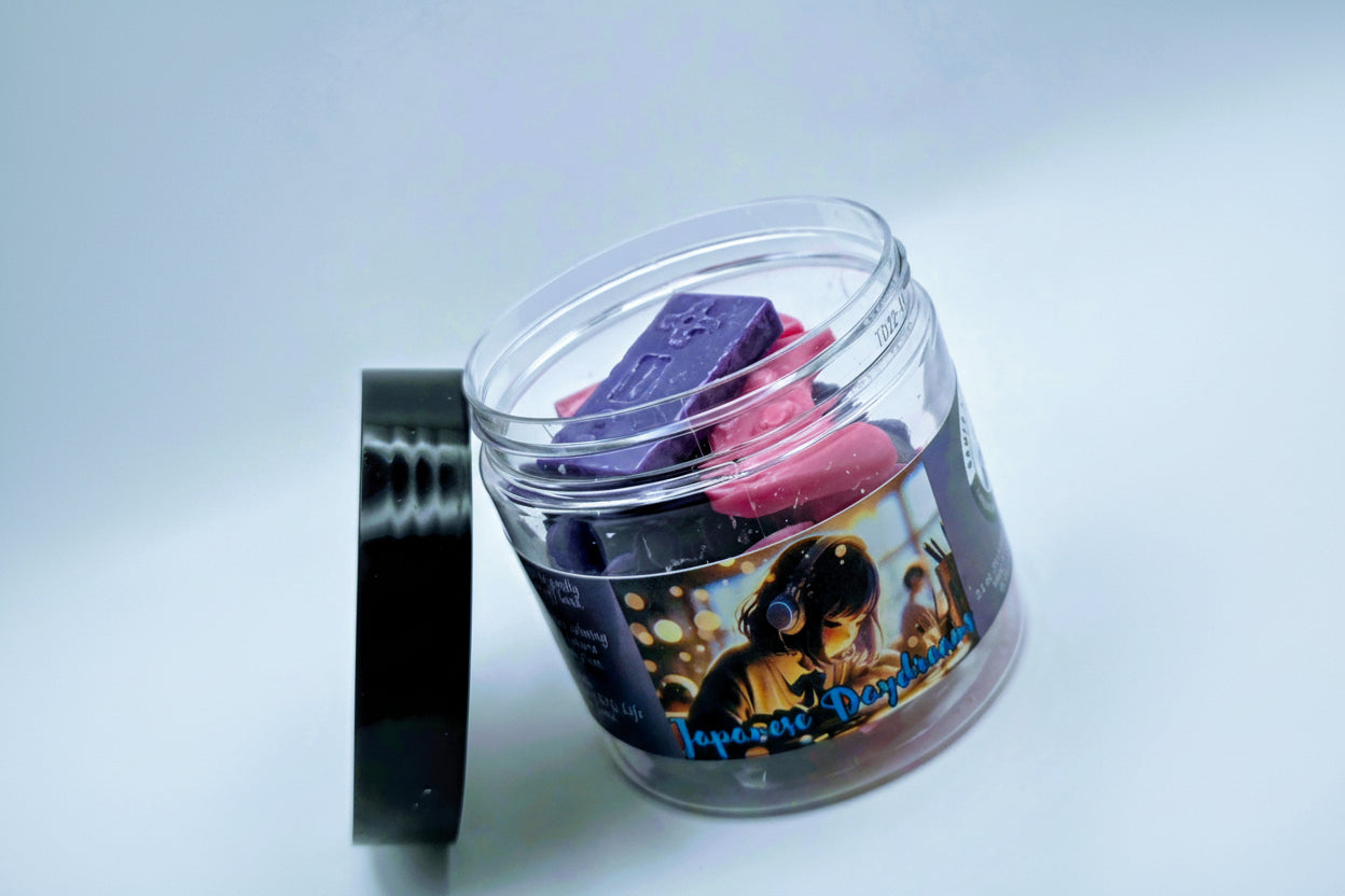 A jar of "Japanese Daydreams" premium wax melts. Each wax melt is shaped like a video game controller, and the melts come in a mixture of purple and pink. The melts come in a clear plastic jar with a black lid and feature a label of a Japanese girl at a school desk, wearing headphones, seeming to lost in thought.