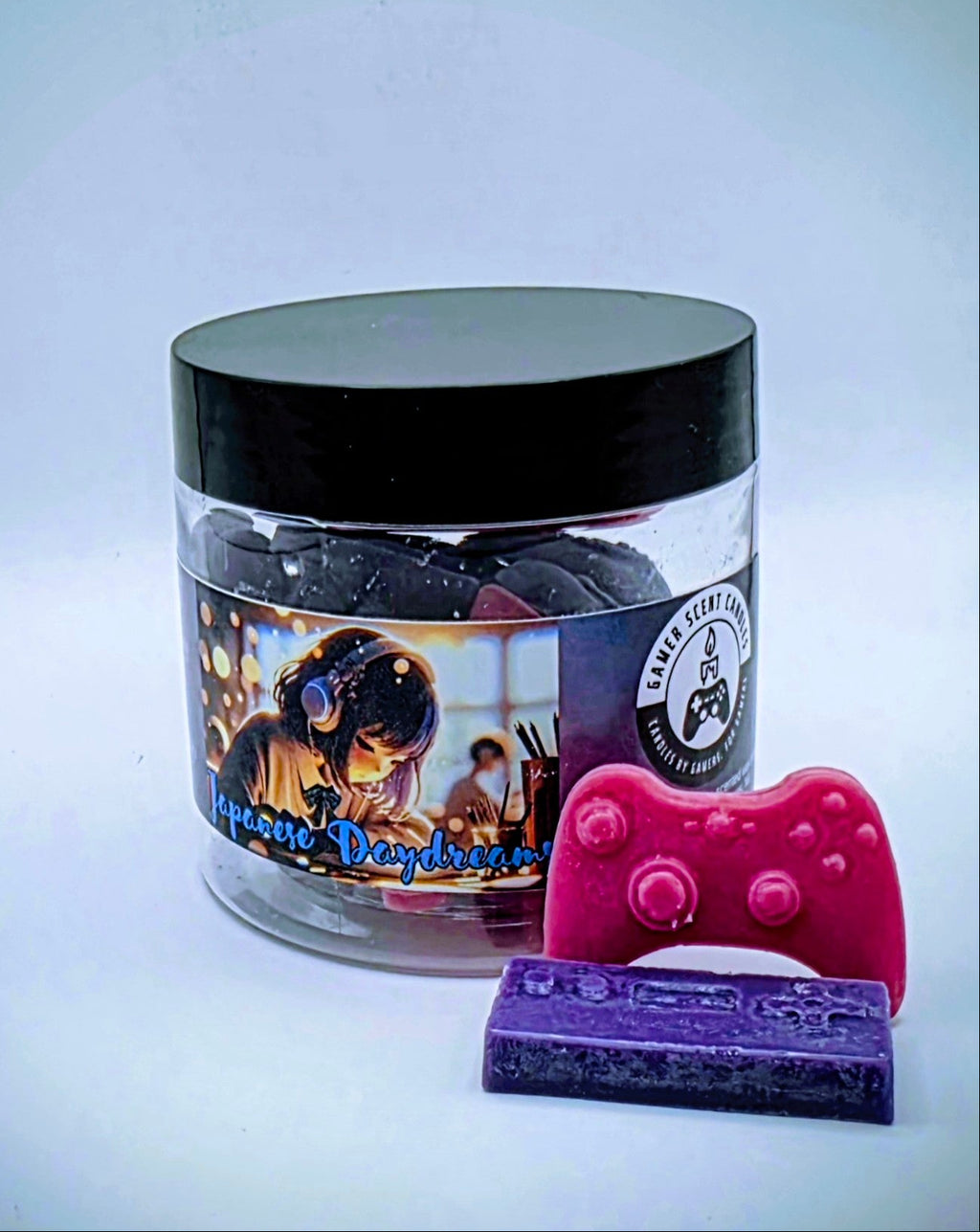 A jar of "Japanese Daydreams" premium wax melts. Each wax melt is shaped like a video game controller, and the melts come in a mixture of purple and pink. The melts come in a clear plastic jar with a black lid and feature a label of a Japanese girl at a school desk, wearing headphones, seeming to lost in thought.