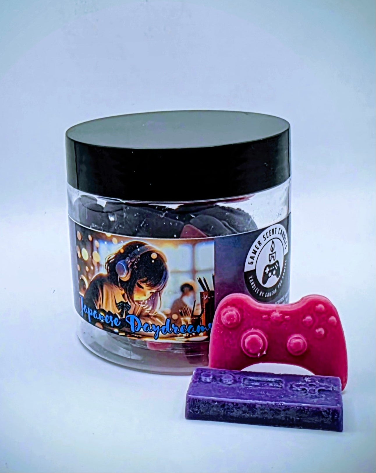 A jar of "Japanese Daydreams" premium wax melts. Each wax melt is shaped like a video game controller, and the melts come in a mixture of purple and pink. The melts come in a clear plastic jar with a black lid and feature a label of a Japanese girl at a school desk, wearing headphones, seeming to lost in thought.