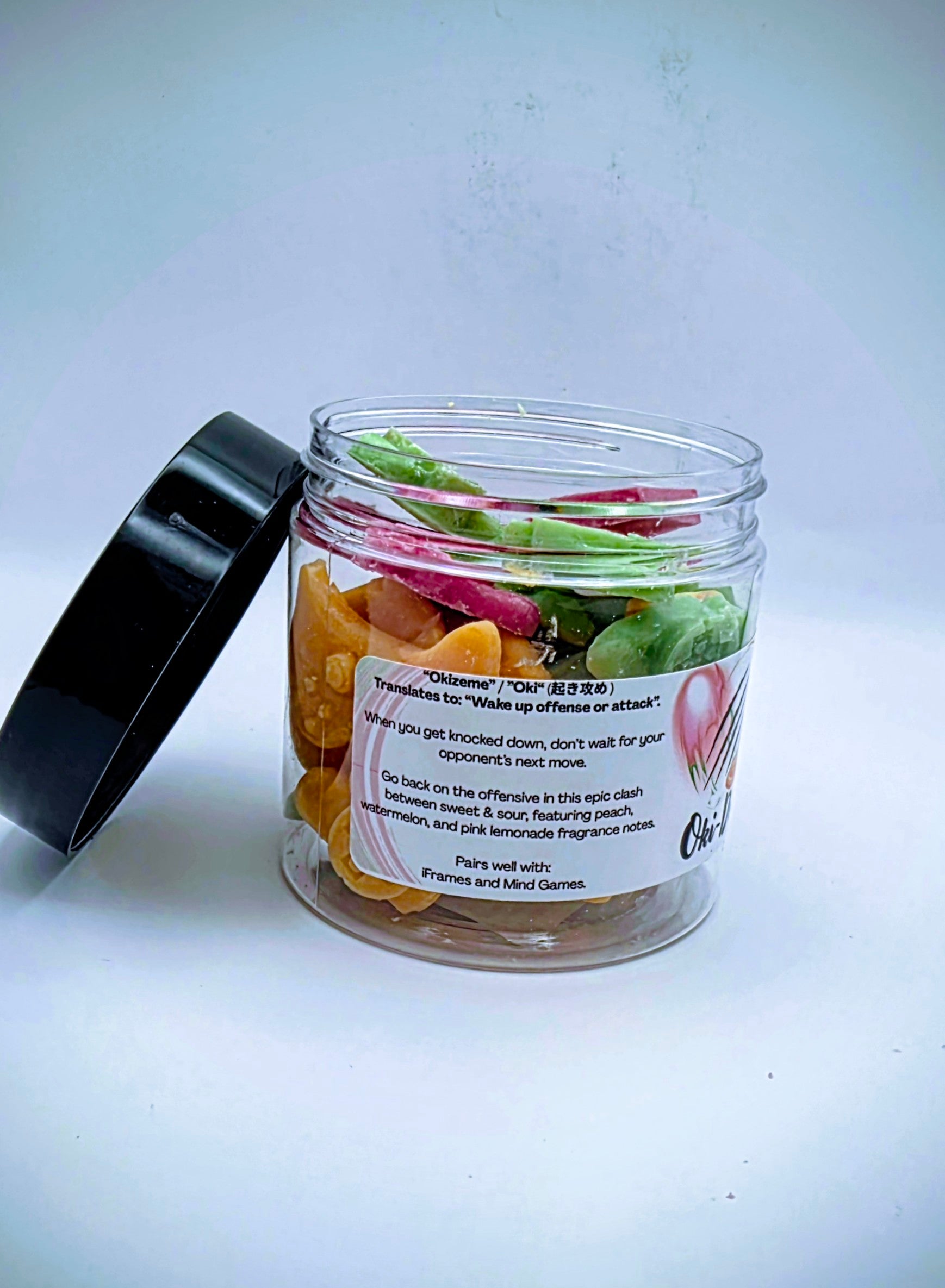 A jar of "Oki-Doki" premium wax melts. The wax melts come in a clear plastic jar, with a white label, featuring imagery reminiscent of fighting games. The wax melts come in a variety of video game controller shapes, and a mix of orange, green, and pink colors.