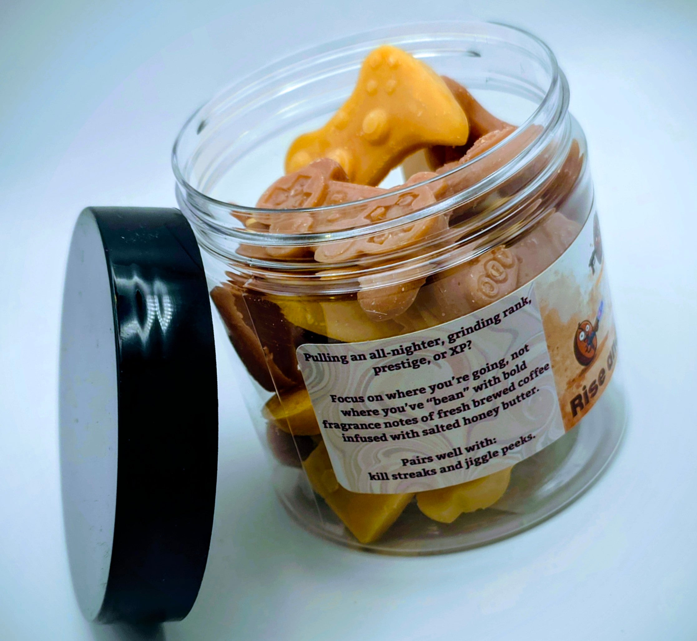 A jar of premium wax melts in an assortment of video game controller shapes. The scent is called "Rise and Grind". The wax melts are a mix of coffee brown and golden cream colors. The label features cartoon coffee beans with weapons.