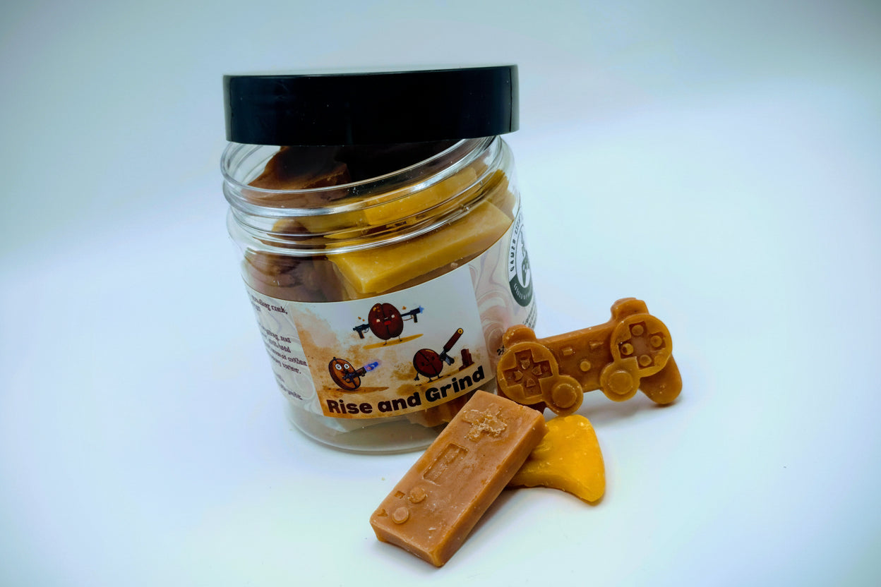 A jar of premium wax melts in an assortment of video game controller shapes. The scent is called "Rise and Grind". The wax melts are a mix of coffee brown and golden cream colors. The label features cartoon coffee beans with weapons.