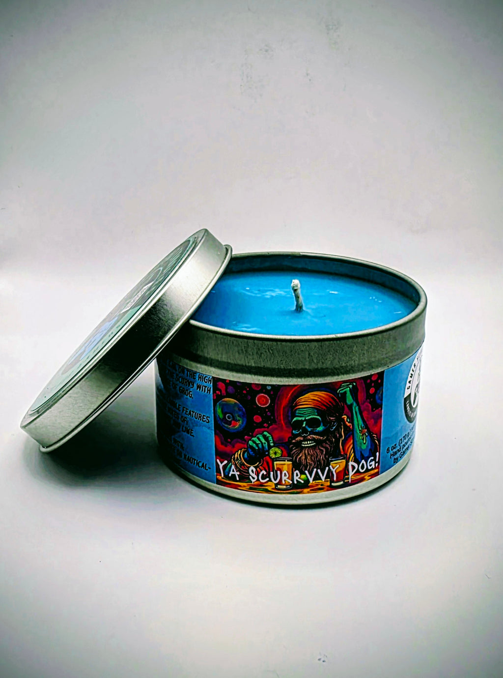 Our "Scurvy Dog" candle in a 6 oz. tin. The candle features spiced coconut and lime fragrance notes. The candle is blue and features a blue label with a psychedelic pirate holding a drink garnished with a lime. Our 6 oz. candles have approximately a 25 hour burn time.
