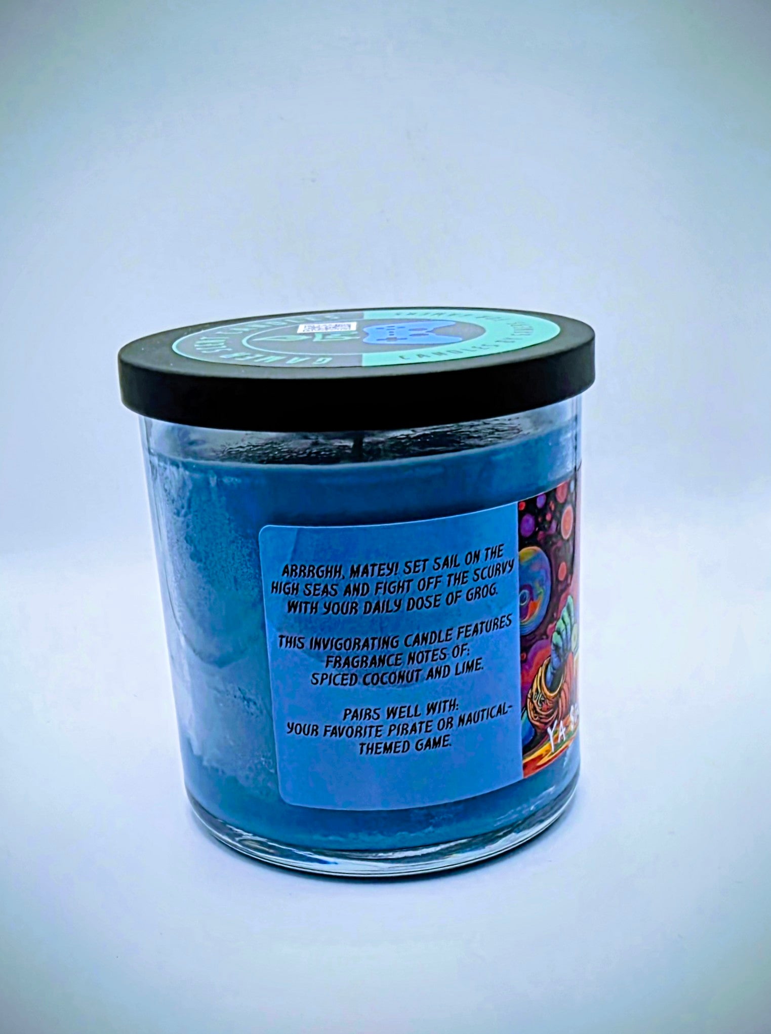 Our "Scurvy Dog" candle in a 9 oz. glass tumbler jar. The candle features spiced coconut and lime fragrance notes. The candle is blue and features a blue label with a psychedelic pirate holding a drink garnished with a lime. Our 9 oz. candles have approximately a 40 hour burn time.