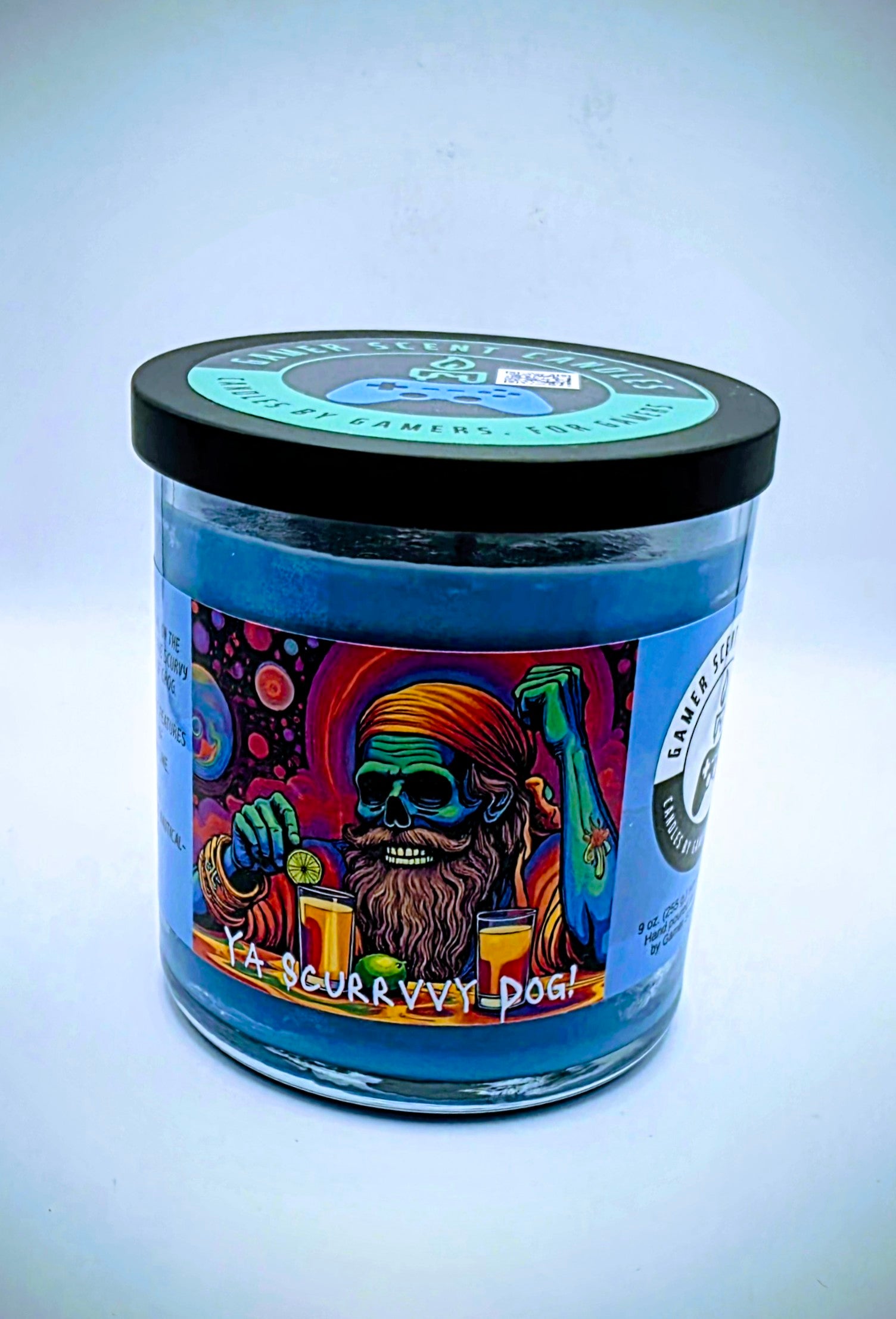 Our "Scurvy Dog" candle in a 9 oz. glass tumbler jar. The candle features spiced coconut and lime fragrance notes. The candle is blue and features a blue label with a psychedelic pirate holding a drink garnished with a lime. Our 9 oz. candles have approximately a 40 hour burn time.