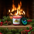 A Holiday Hearth 9-ounce tumbler jar candle, sitting in front of a holiday fireplace, surrounded by mistletoe and pinecones. Front view. Lid off. Lit.  Video game candle. Perfect gift for gamers. Premium soy wax scented candle.