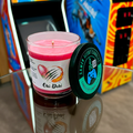 Oki-Doki 9-ounce tumbler jar. On a table, in front of an arcade cabinet. Front view. Lid off.  Video game candle. Perfect gift for gamers. Premium soy wax scented candle.